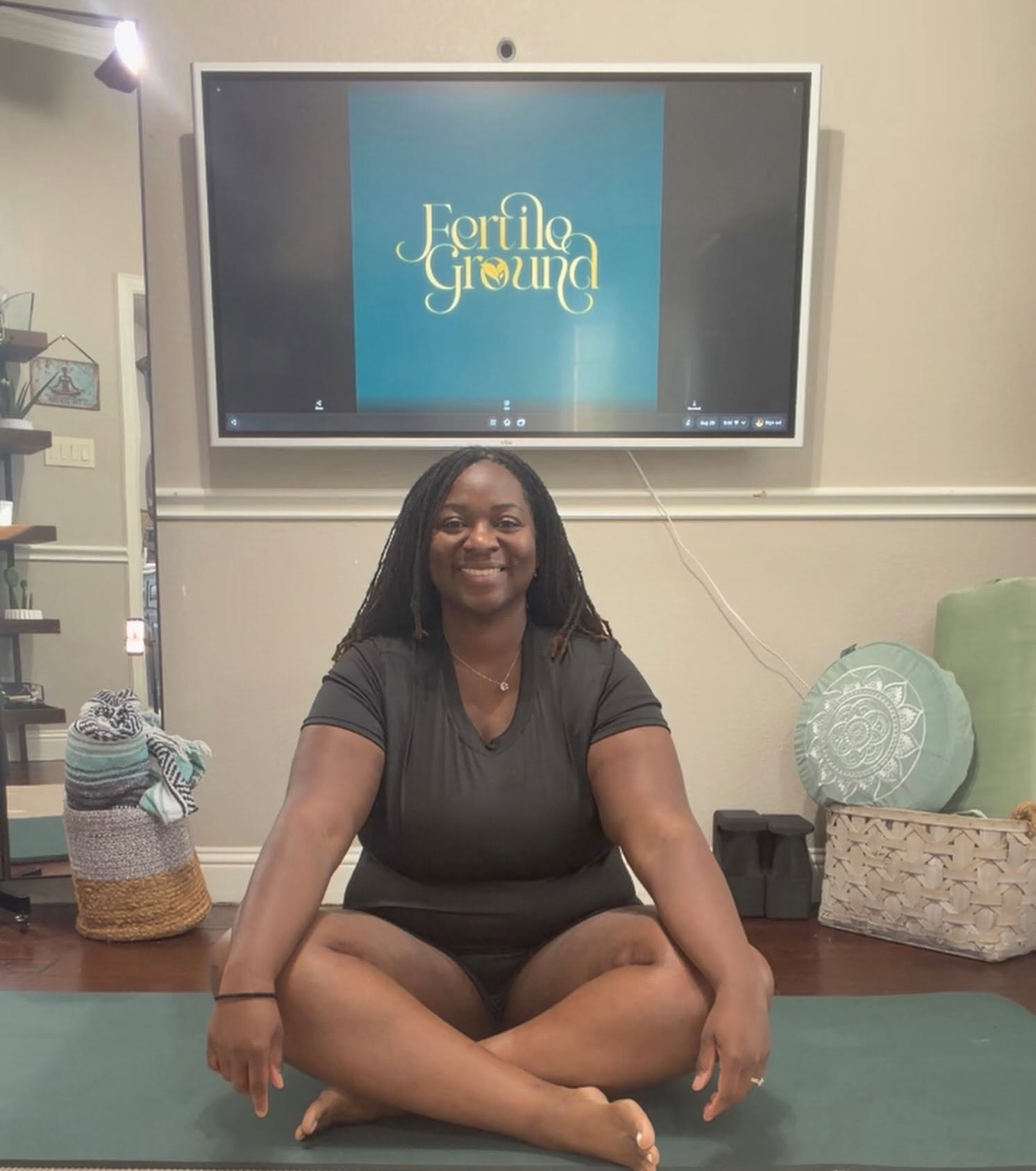 Quisha seated cross-legged on a yoga mat in her home practice space with the Fertile Ground logo on a screen behind her