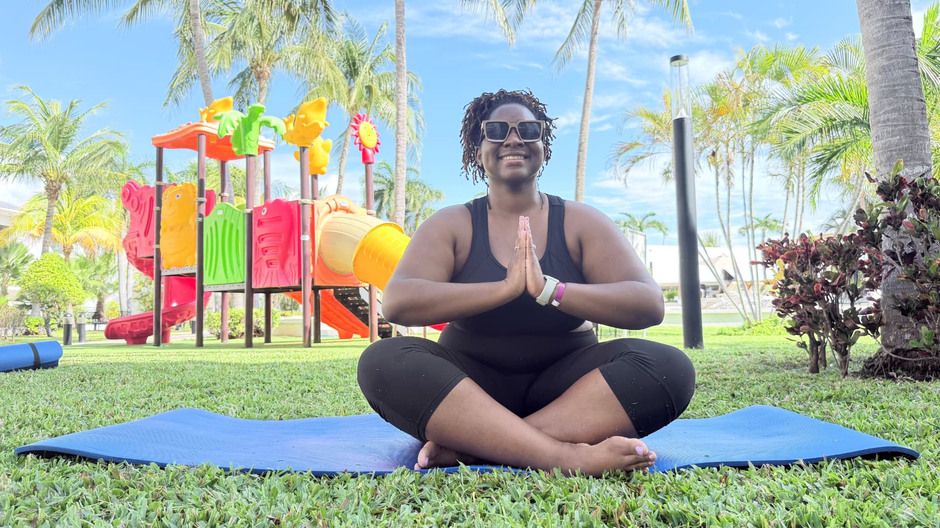 Quisha seated cross-legged on a yoga mat with her hands together outdoors