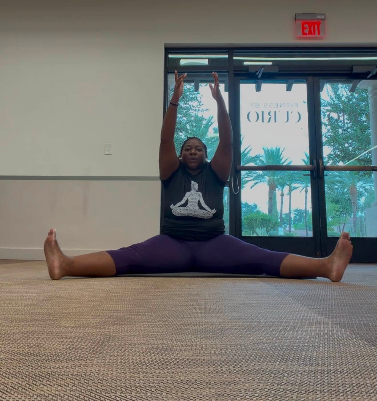 Quisha demonstrating a seated wide-legged stretch with both arms raised